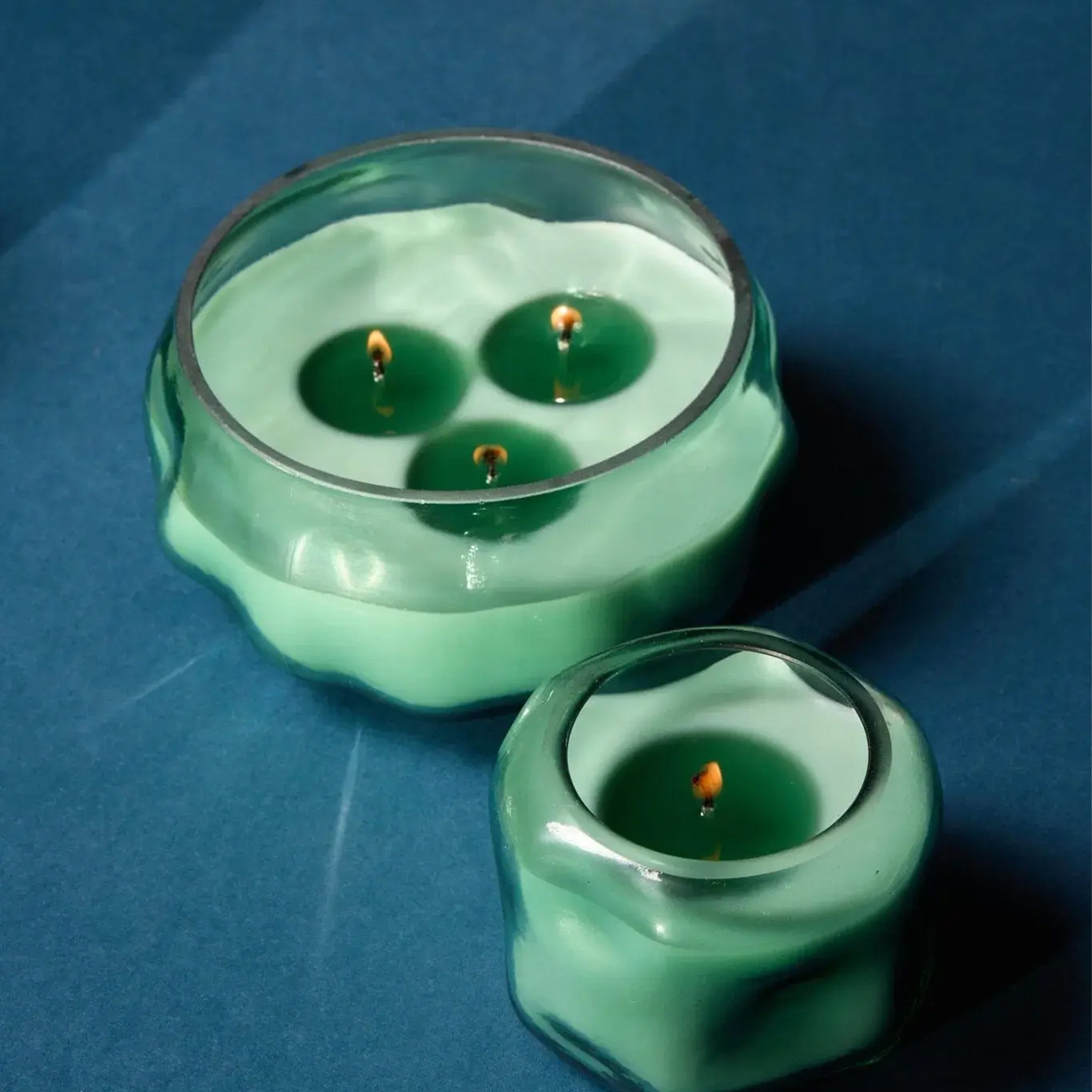 A large scalloped green glass candle with three flames sits beside a smaller matching candle, both glowing softly against a rich blue background.