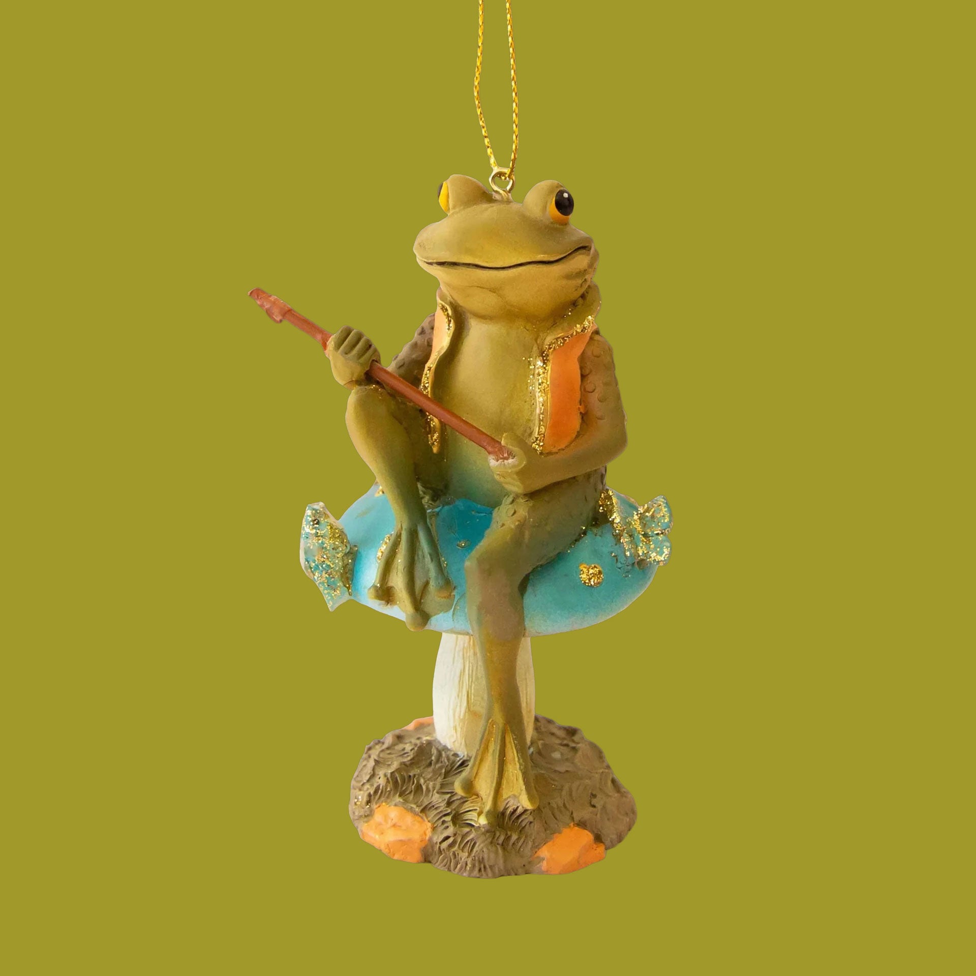 Ornament of a green frog with an orange vest on, sitting on top of a blue mushroom. The frog is holding a fishing pole and there is a blue fish with gold sparkles. 