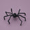 A black fuzzy spider decoration placed on a solid light purple background.