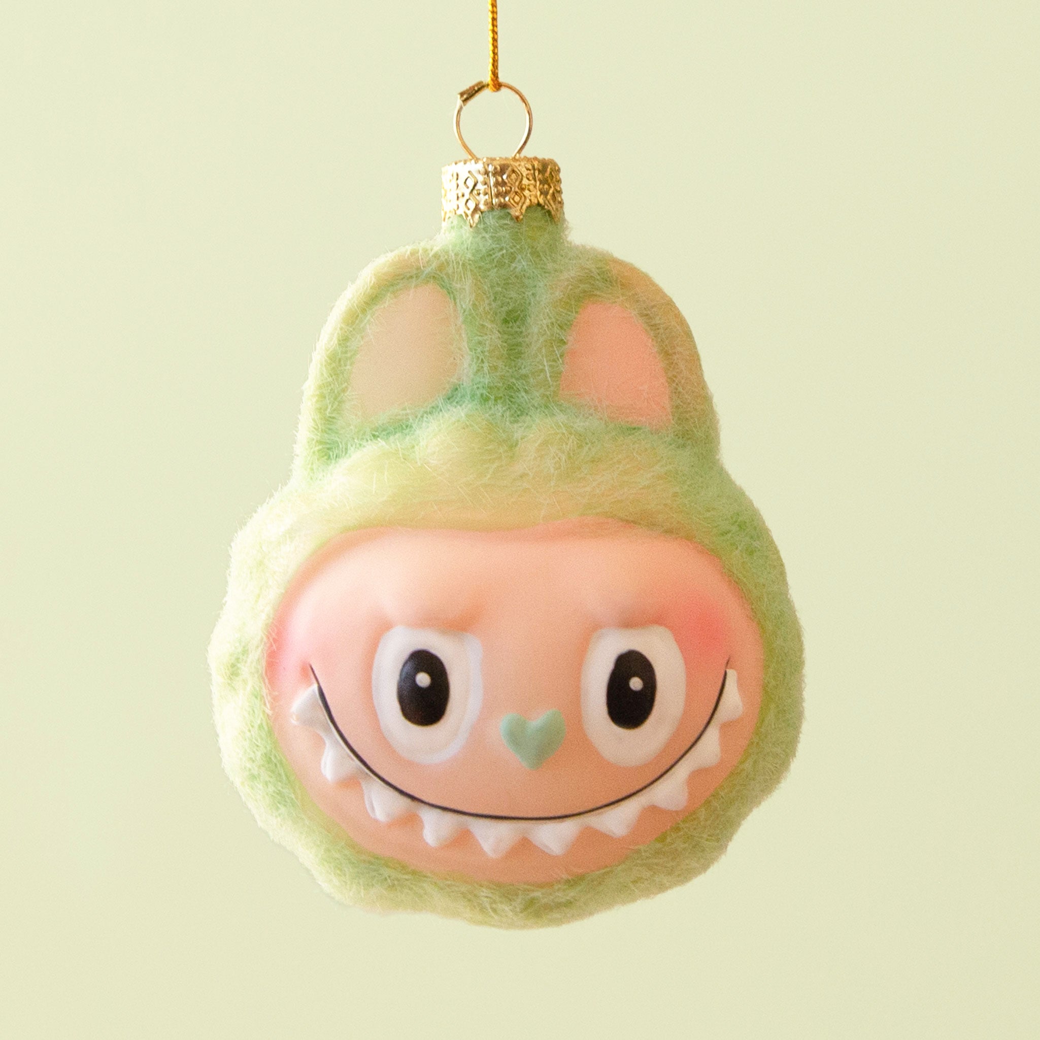 A small green fuzzy monster ornament with closed eyes, a tiny orange nose, and a gentle smile hangs from a gold cap and yellow string against a pale green background.