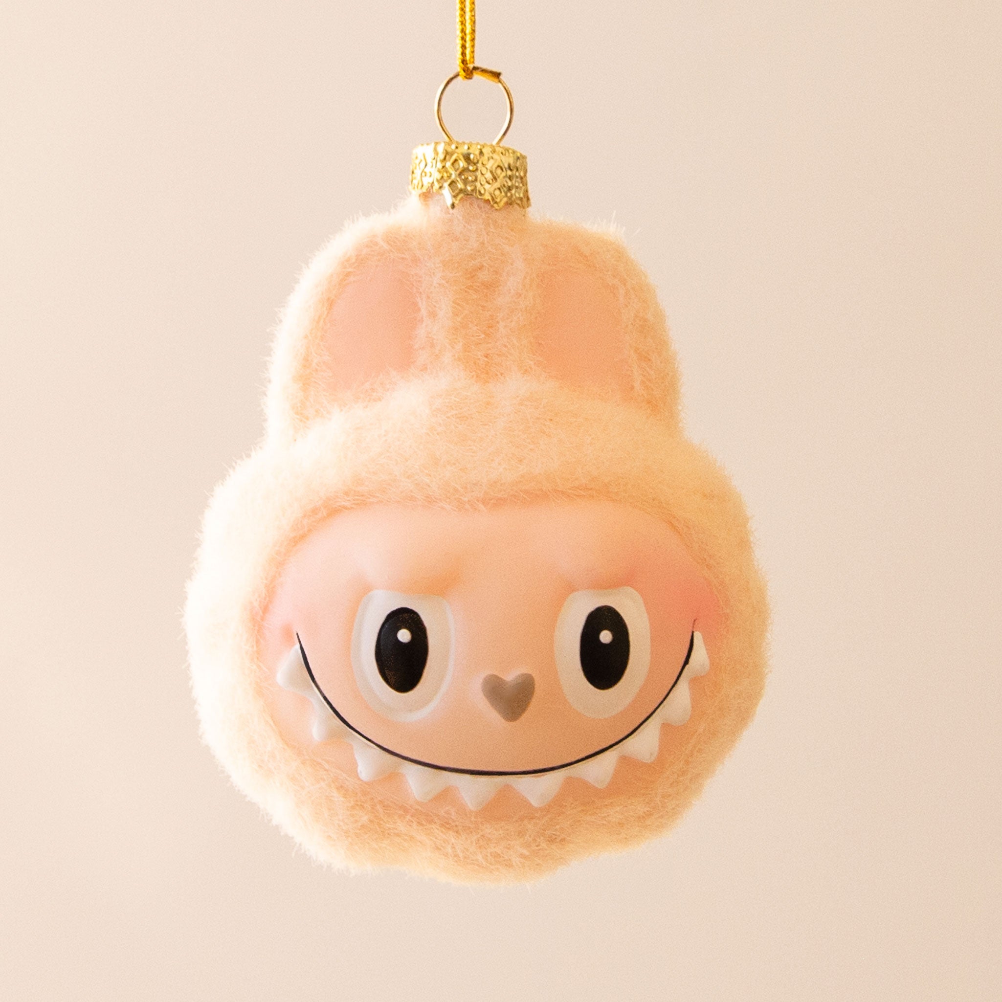 A small ivory fuzzy monster ornament with closed eyes, a tiny orange nose, and a gentle smile hangs from a gold cap and yellow string against a pale cream background.