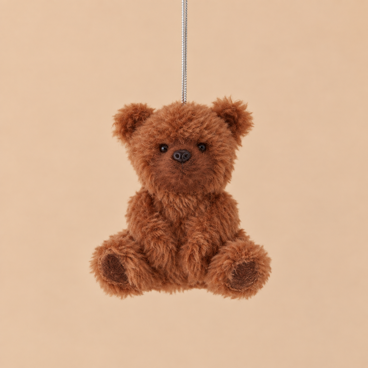 A small brown plush teddy bear ornament hanging from a thin silver string against a solid warm beige background.