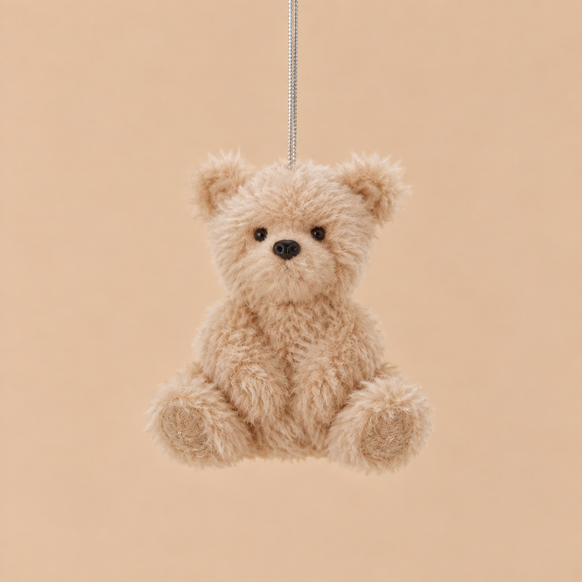 A small plush teddy bear ornament in light beige faux fur, hanging from a thin silver string against a solid warm beige background.