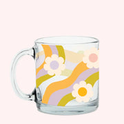 On a light pink background is a clear glass mug with a multicolored wavy print and white daisies scattered. 