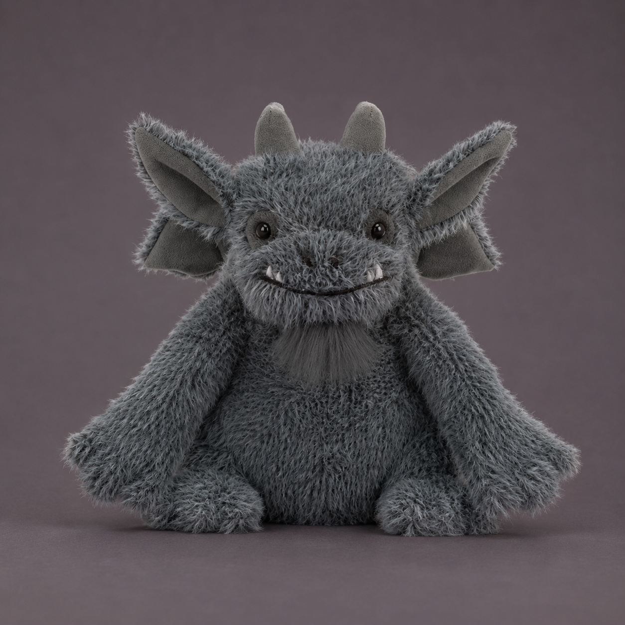 A gray plush gargoyle toy with large pointed ears, small horns, and a stitched smile, photographed against a plain mauve-gray studio background.