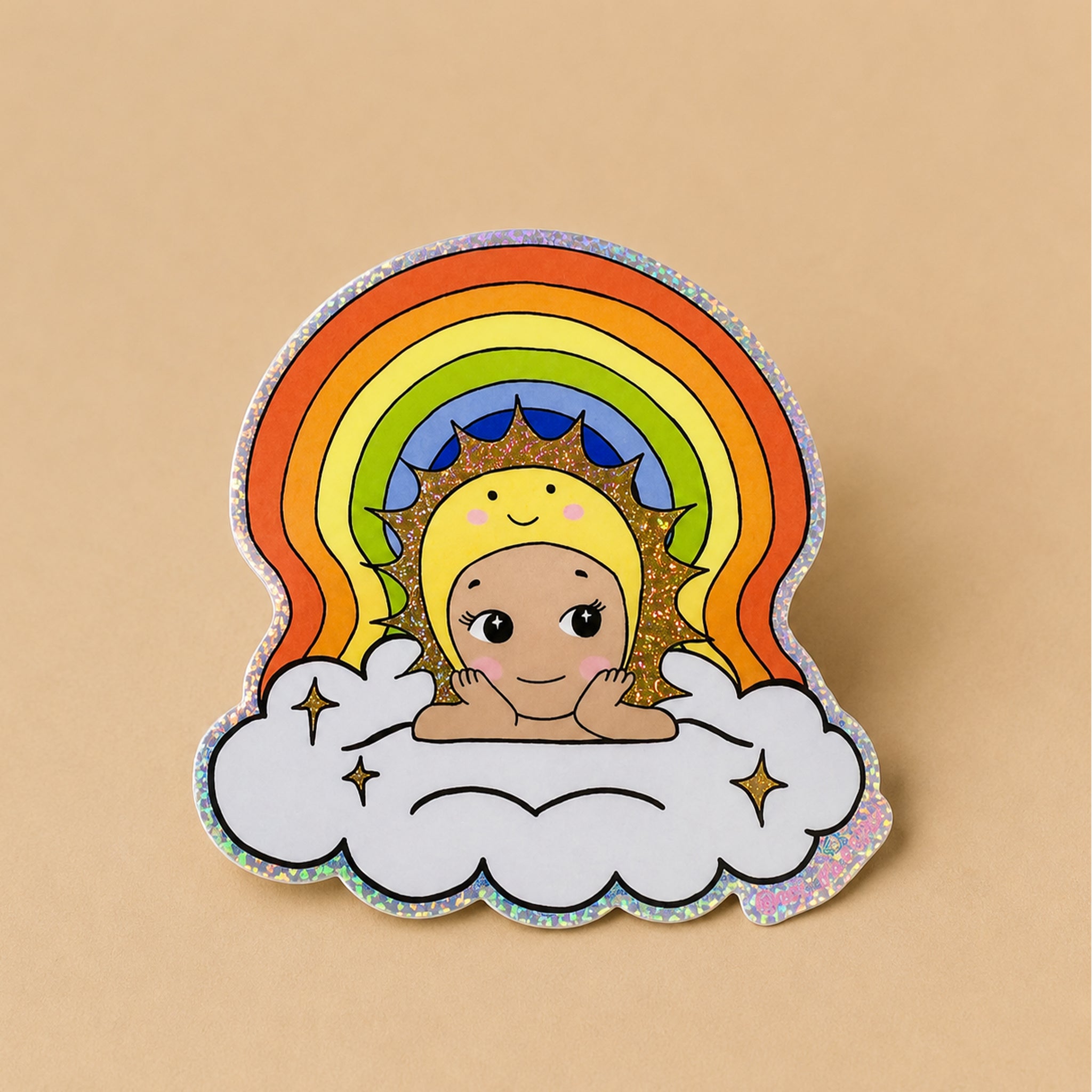 A die-cut enamel pin featuring a smiling baby sun peeking out from behind a glittery rainbow and white cloud, set against a solid beige background.