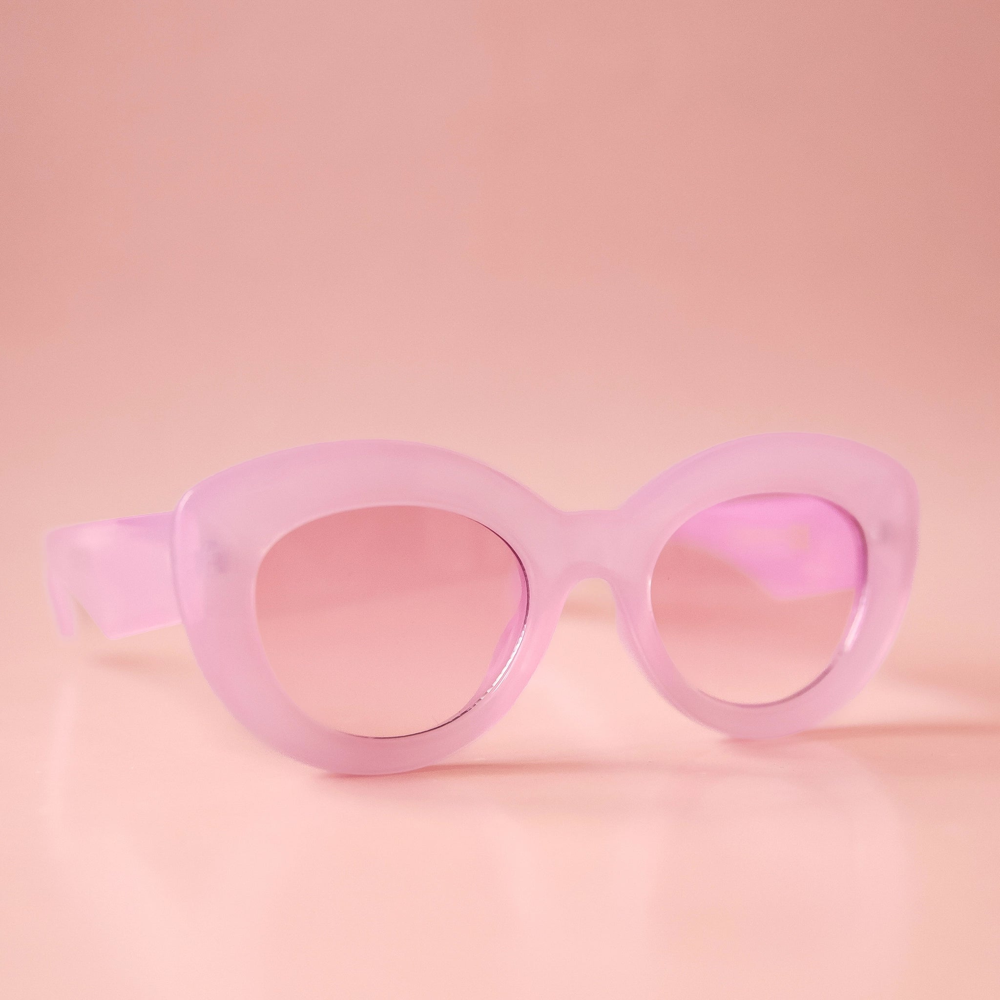 On a peachy background is a pair of light purple sunglasses with a rounded, slightly cat eye shape and a light pink/peach lens.