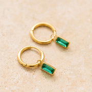 On a tan background is gold hoop earrings with an emerald cut emerald stone hanging from it. 