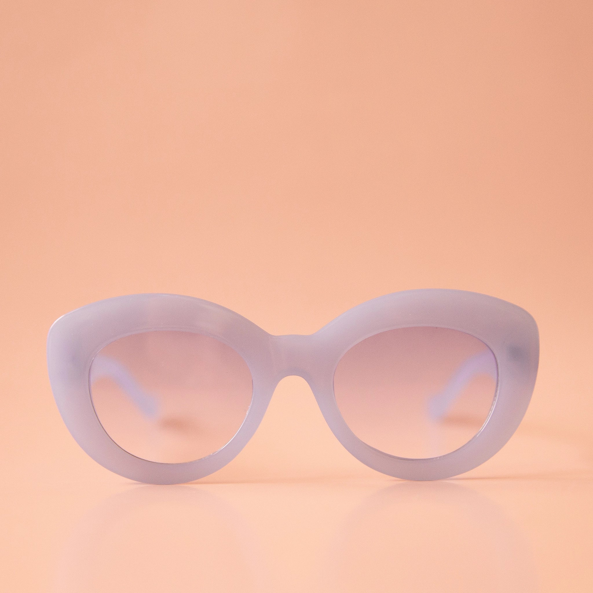 On a peachy background is a pair of light blue sunglasses with a rounded shape and a slight cat eye corner. The lenses are also a light blue shade