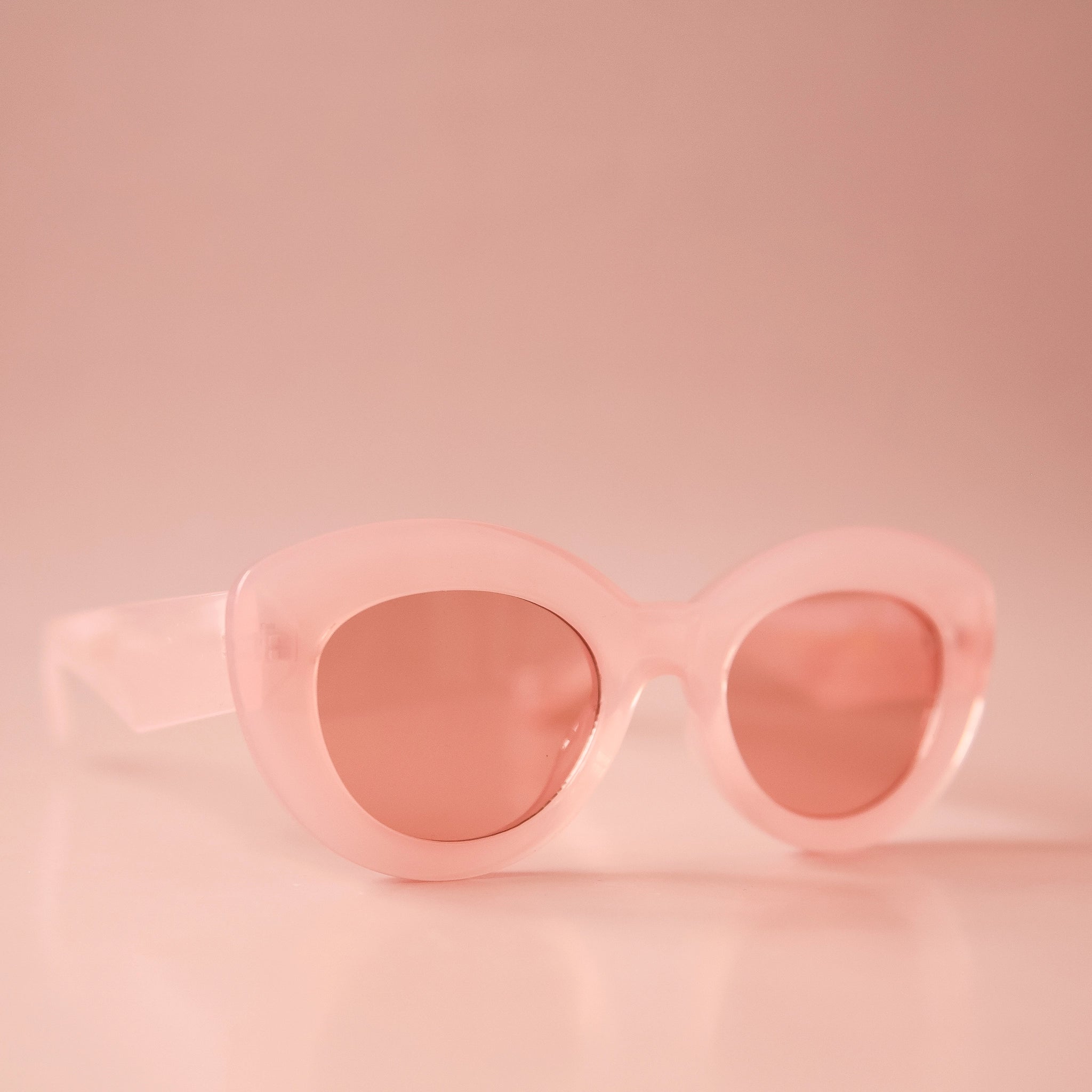 On a light peachy background is a pink pair of sunglasses with a rounded shape and a slight cateye at the corner along with a peachy lens. 