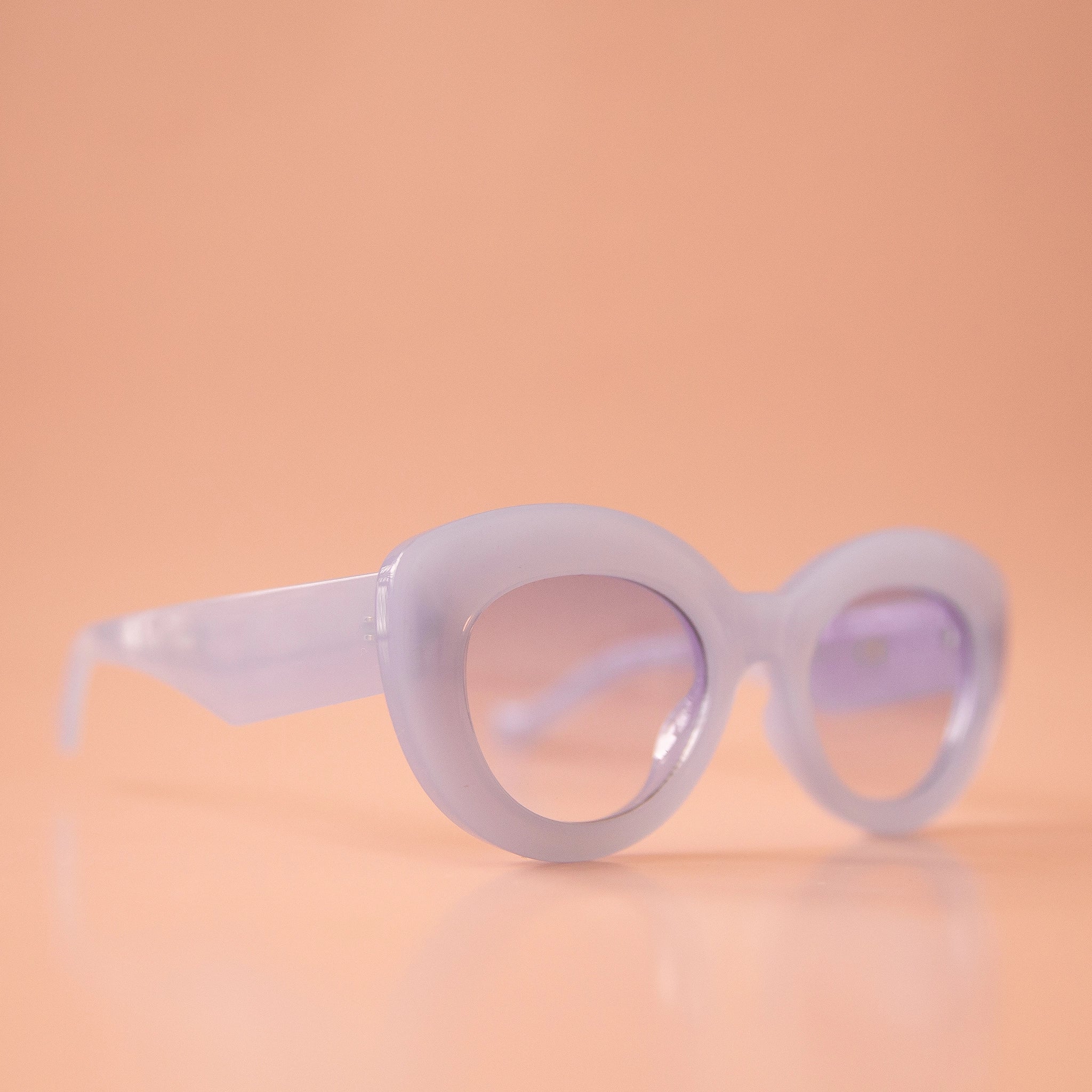 On a peachy background is a pair of light blue sunglasses with a rounded shape and a slight cat eye corner. The lenses are also a light blue shade. 