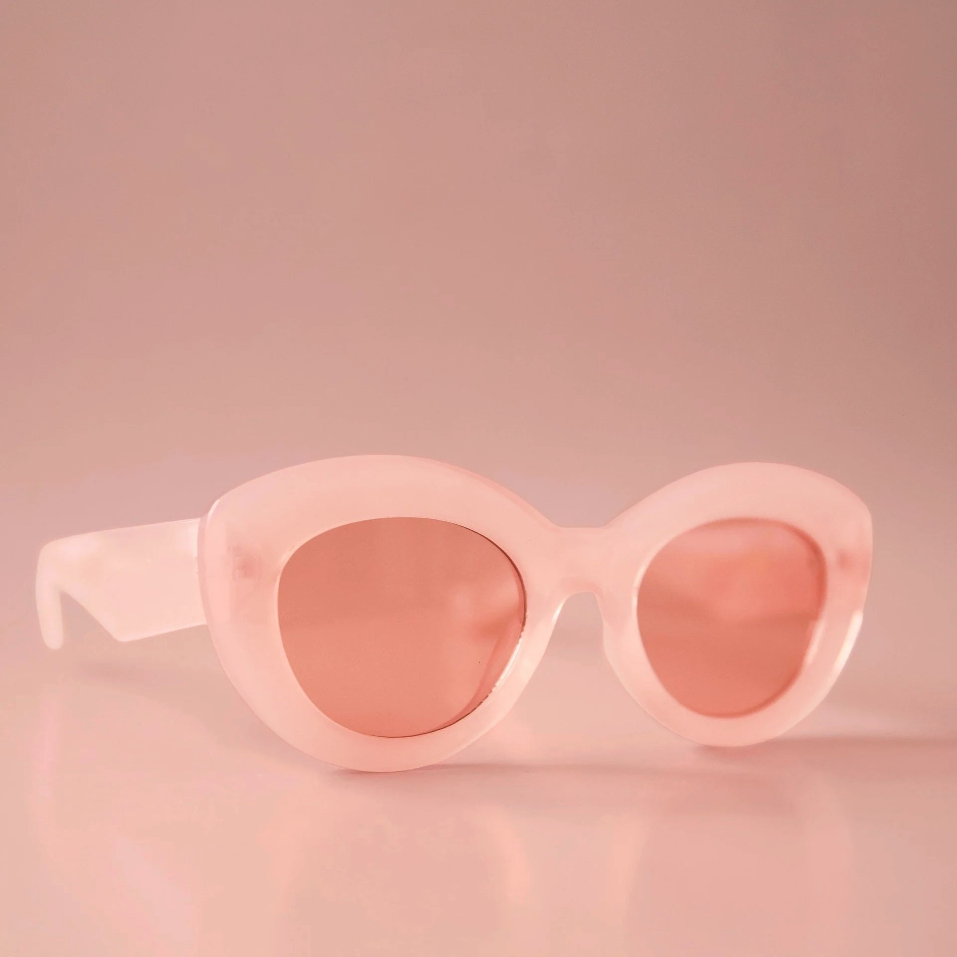 On a light peachy background is a pink pair of sunglasses with a rounded shape and a slight cateye at the corner along with a peachy lens. 