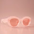 On a light peachy background is a pink pair of sunglasses with a rounded shape and a slight cateye at the corner along with a peachy lens. 