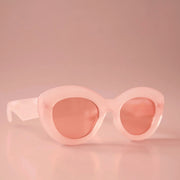 On a light peachy background is a pink pair of sunglasses with a rounded shape and a slight cateye at the corner along with a peachy lens. 