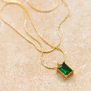 On a tan background is a gold chain necklace with an emerald green and emerald cut stone in the center. 