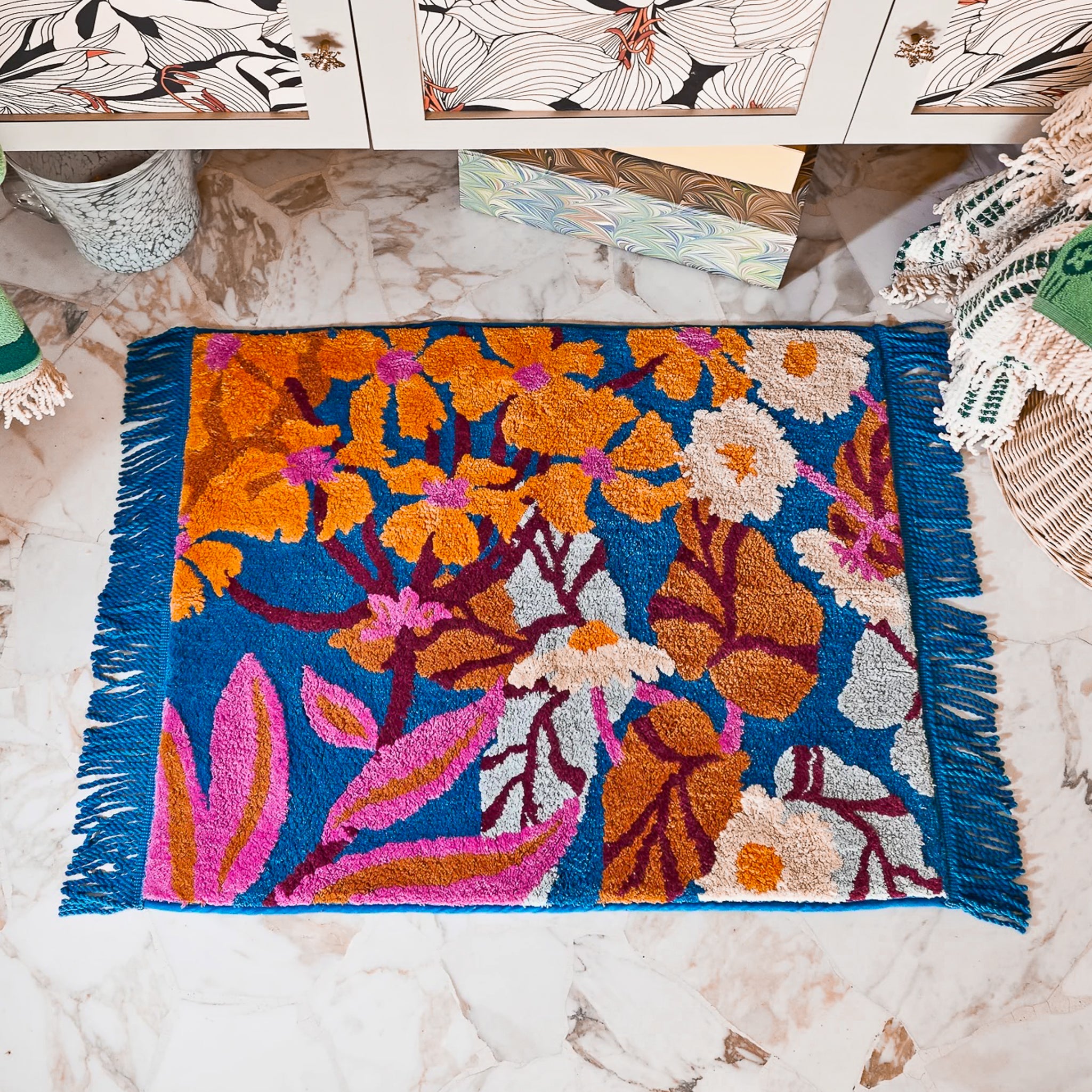Blue bath mat with blue fringe, with orange, pink and white flowers.