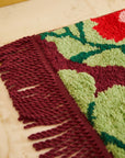 A maroon, pink, red and green bath mat with fringe details on two of the ends. 