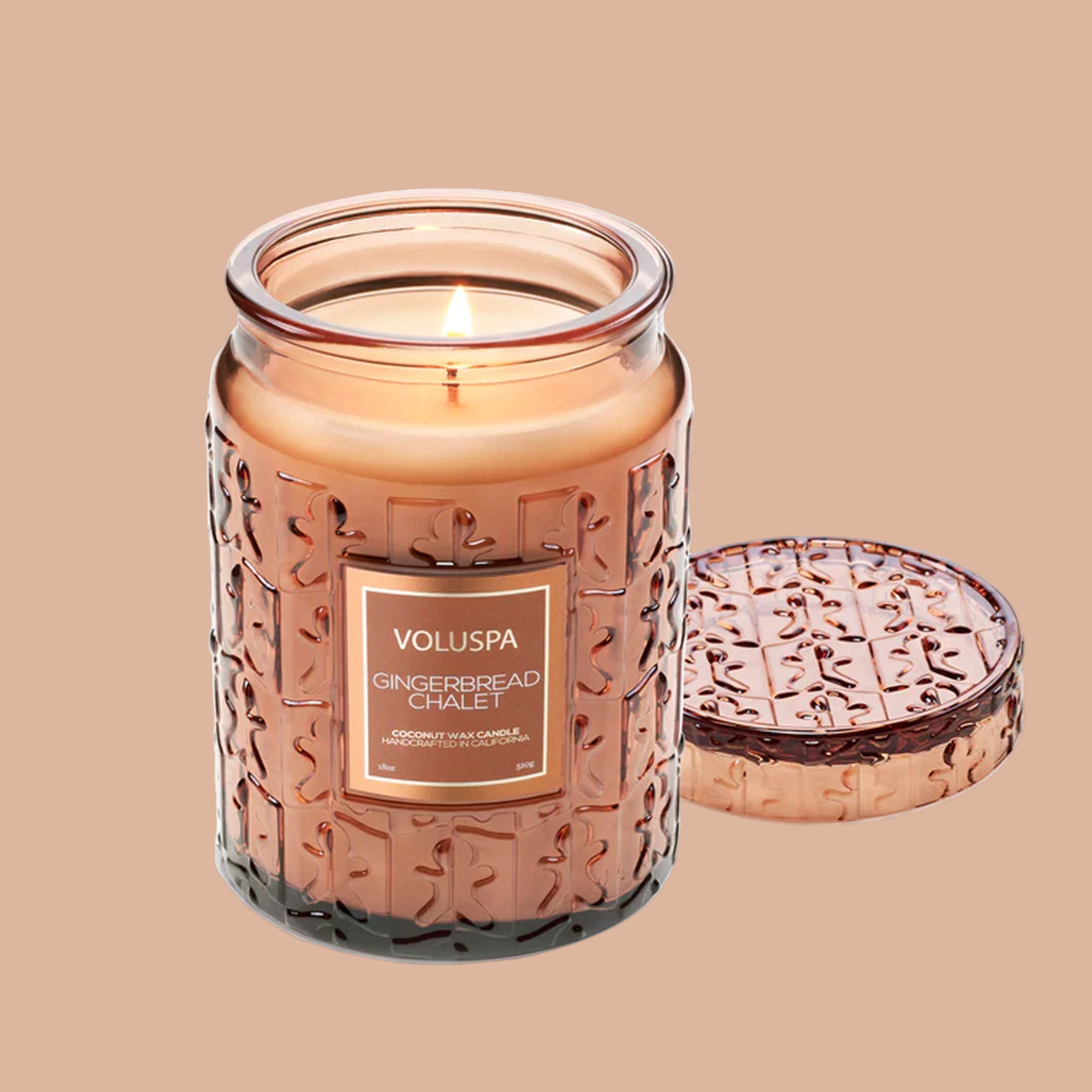 A brown translucent decorative glass jar candle with raised gingerbread shaped pattern and a label in the center that reads, &#39;Voluspa Gingerbread Chalet&#39;. 