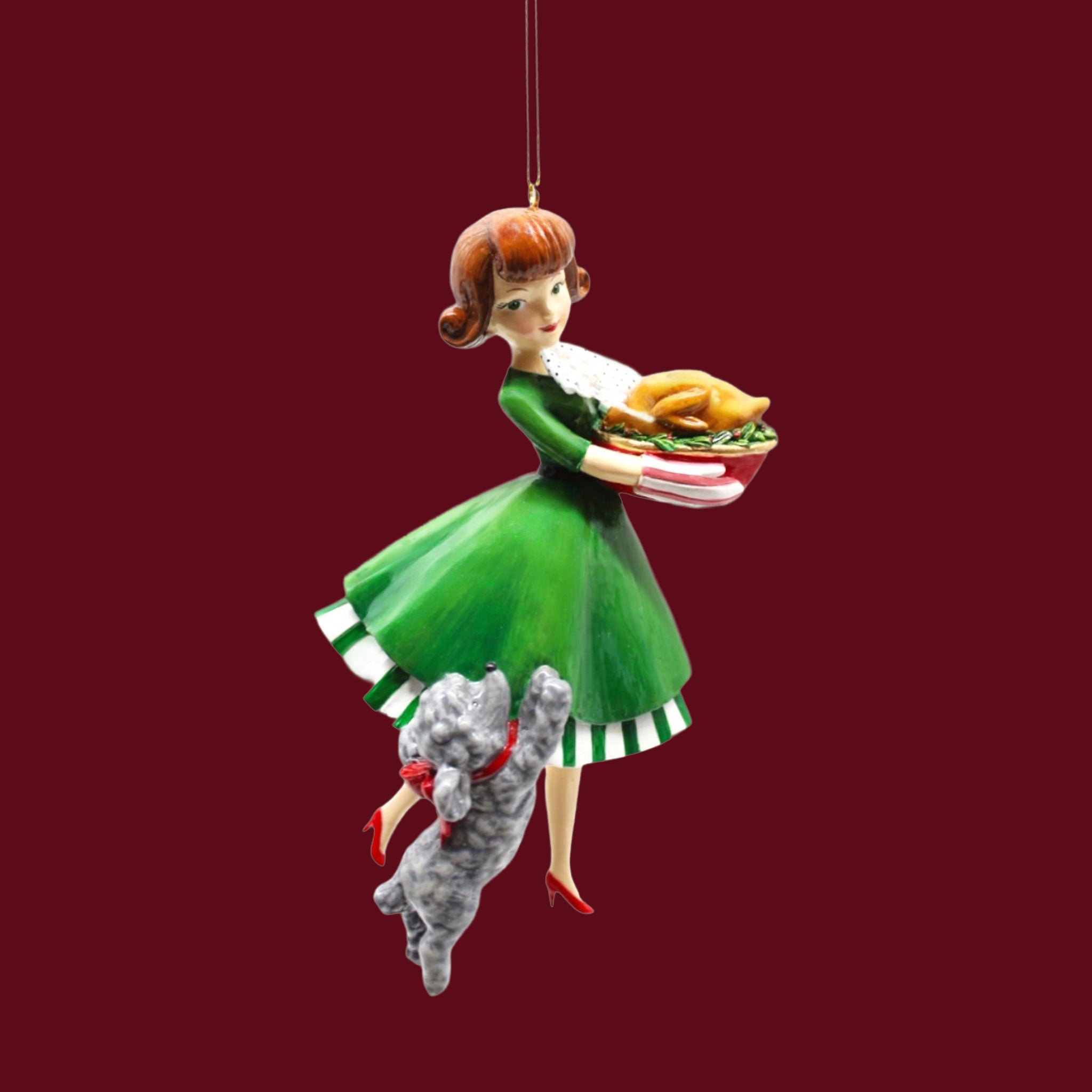 A vintage inspired Christmas ornament. A retro style woman with brown hair and a green dress holds a turkey with a gray dog barking below her.