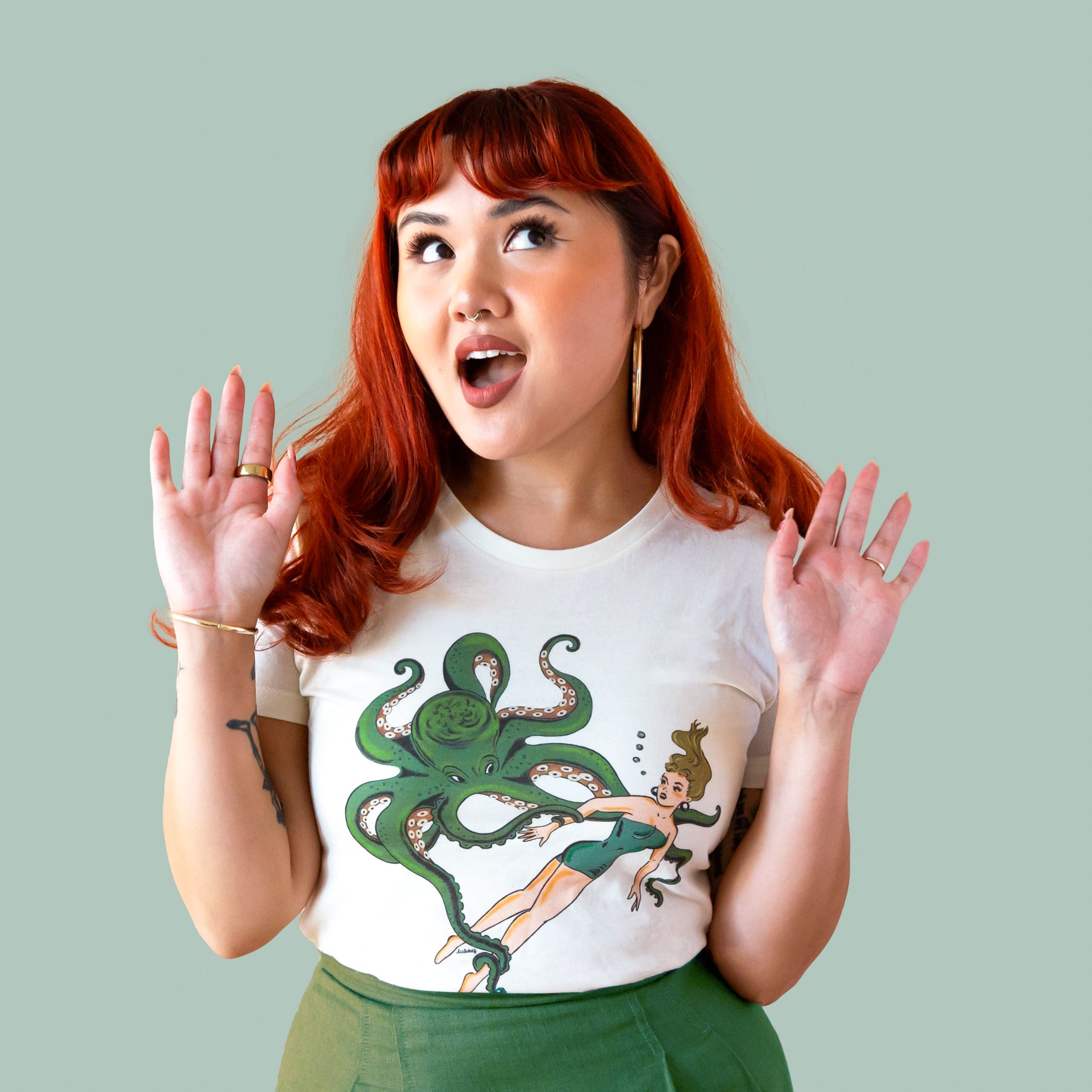 Model with red hair is wearing a white tee shirt detailing a green octopus holding on to a girl swimming. 