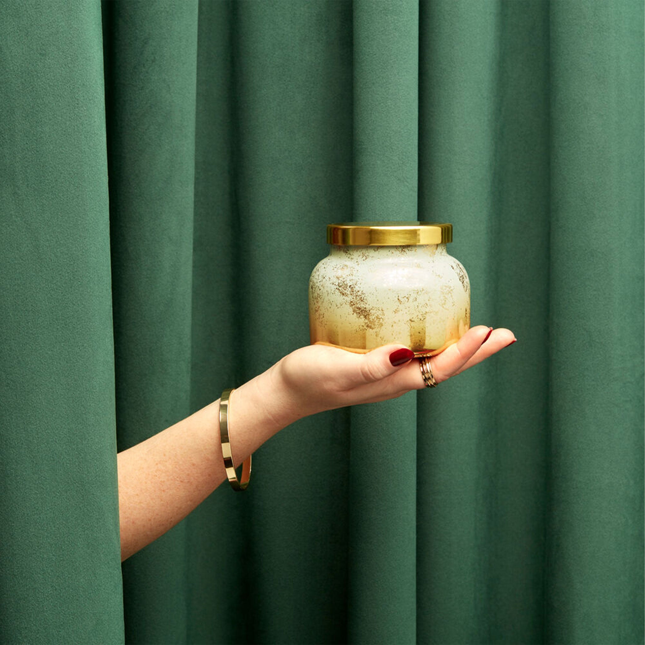A model&#39;s hand holding a white and gold glass jarred candle with a gold lid that reads, &quot;capriBLUE Volcano&quot;