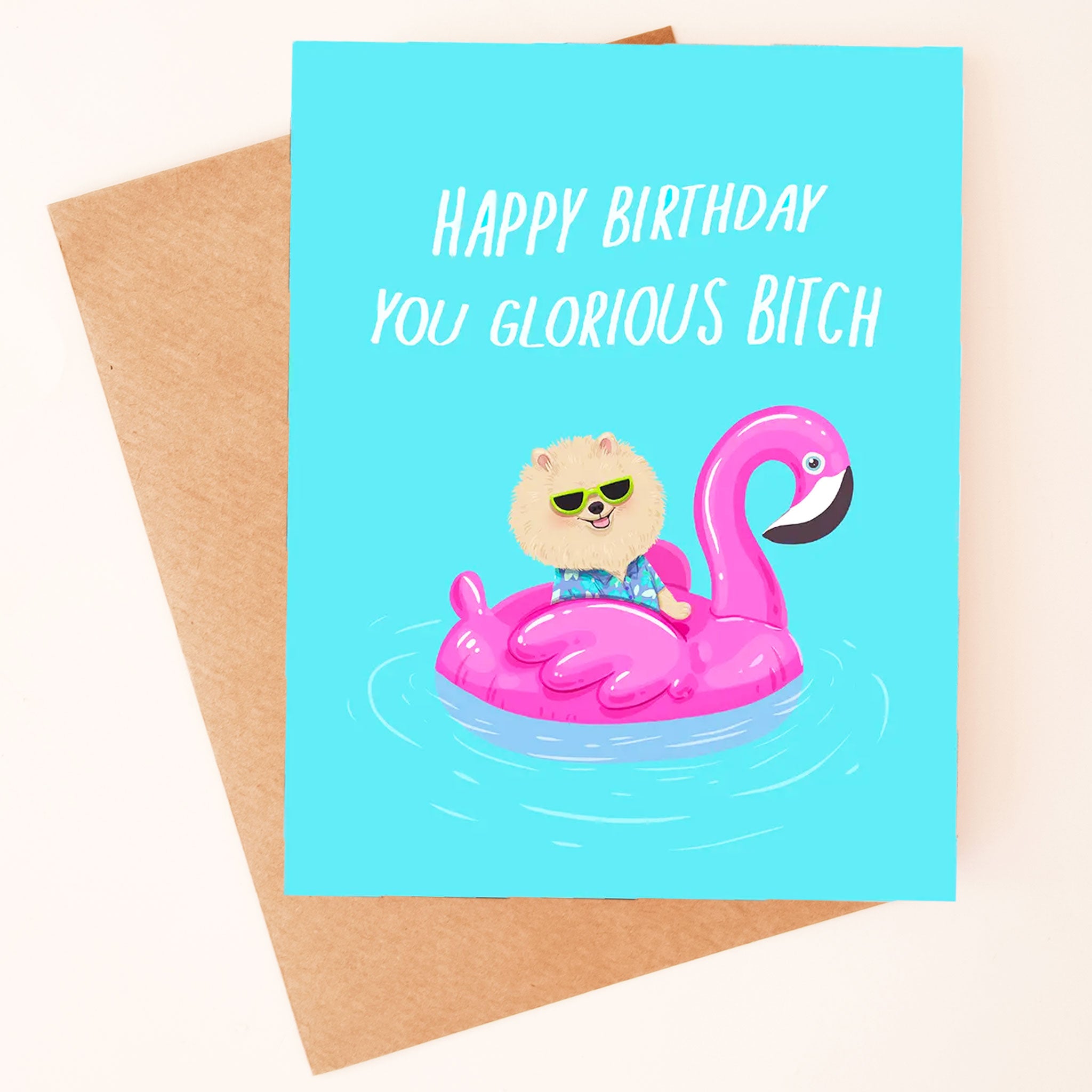 On a neutral background is a bright blue card with an illustration of a Pomeranian in sunglasses on a pink pool float and white text that reads, "Happy Birthday You Glorious Bitch". 