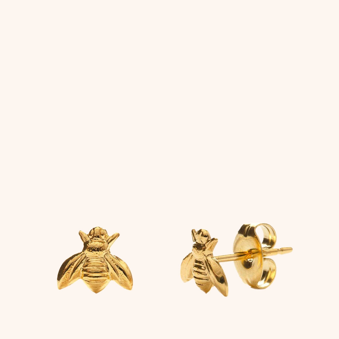 On a cream background is a pair of gold stud earrings in the shape of a honey bee. 
