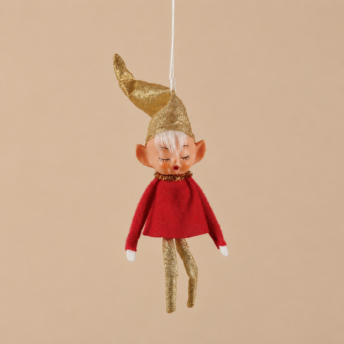 A small hanging elf ornament with a glittery gold pointed hat, pale skin, closed eyes, a red dress, and glittery gold legs, suspended against a plain beige background.