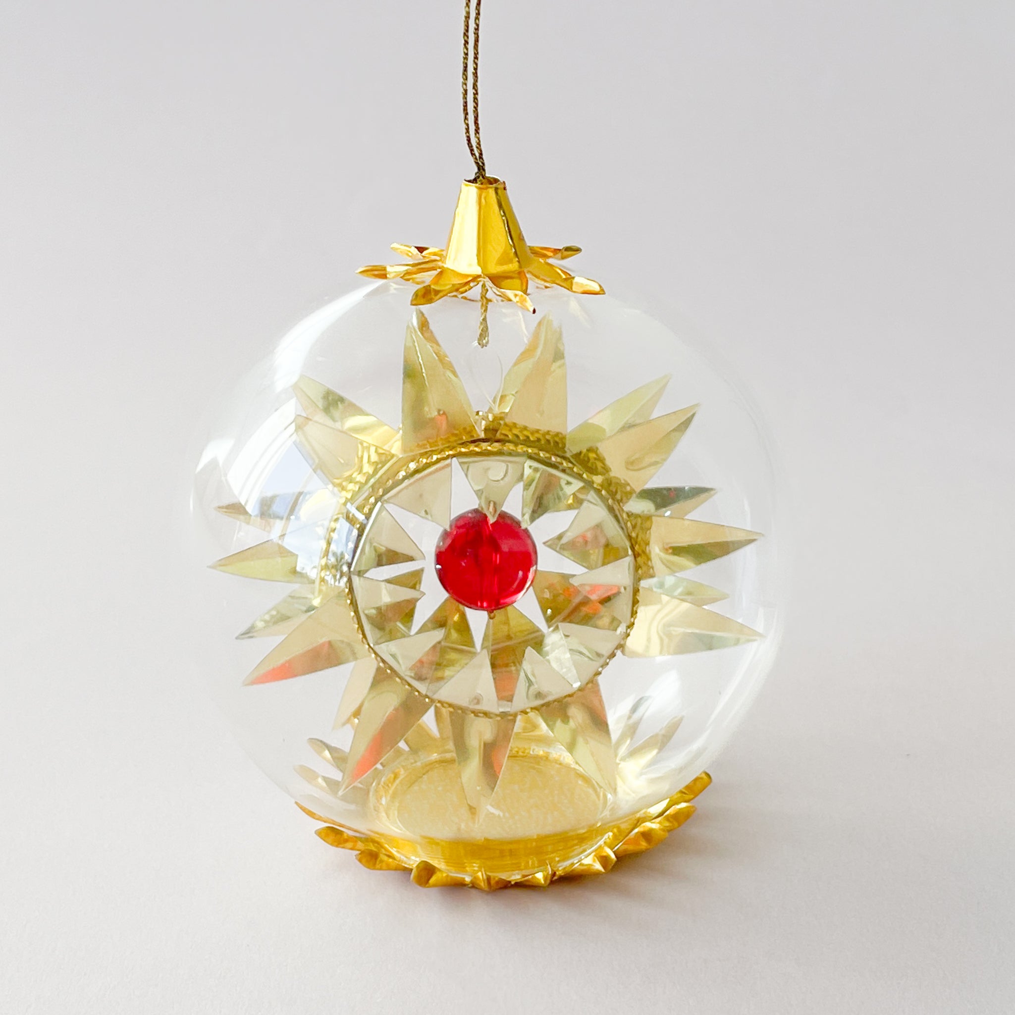 A clear round glass ornament hangs on a white background, featuring a metallic gold starburst design inside with a small red jewel at the center, and a gold cap and base.