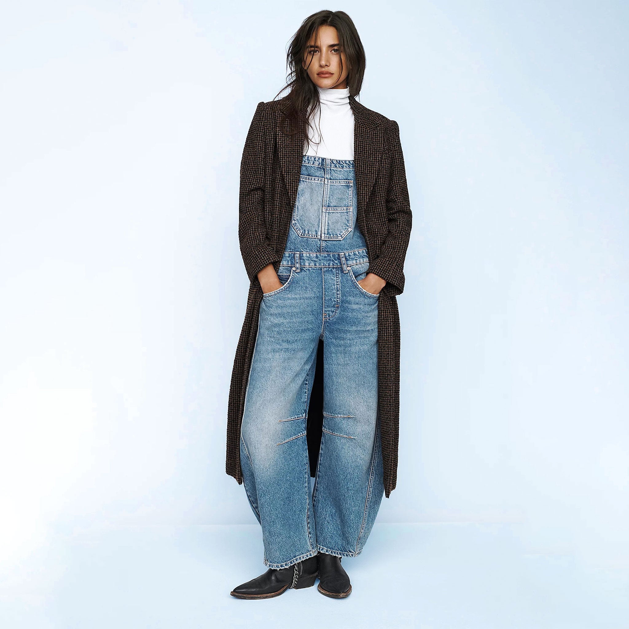 A brown haired model wears a pair of denim overalls and a long tweed coat.