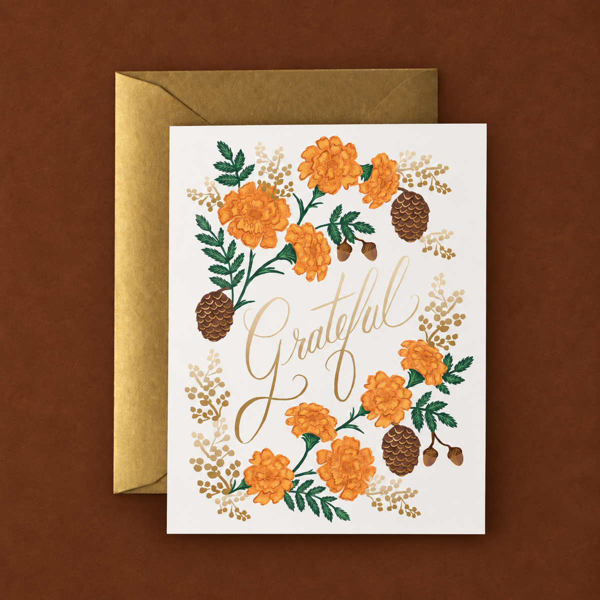 A white greeting card with orange florals and brown pinecones and a gold envelope on a dark brown ground.  The card reads "grateful" in gold.
