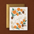 A white greeting card with orange florals and brown pinecones and a gold envelope on a dark brown ground.  The card reads "grateful" in gold.