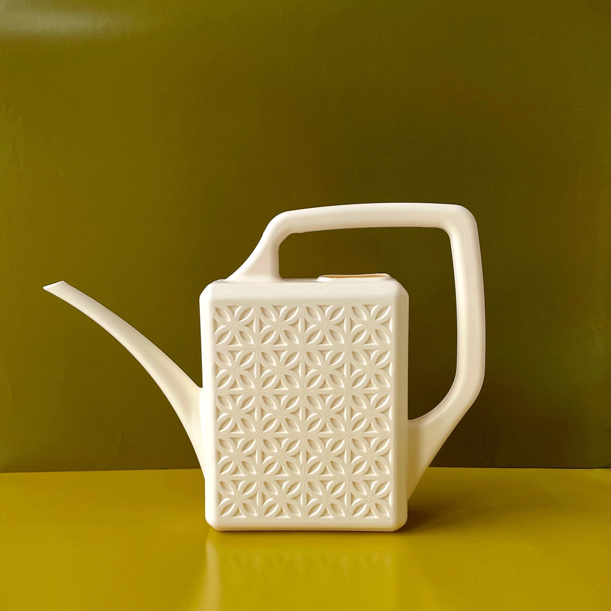 An ivory plastic watering can with a narrow spout and square handle and a rectangle breeze block design on the sides. The background is an olive green hue.
