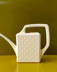 An ivory plastic watering can with a narrow spout and square handle and a rectangle breeze block design on the sides. The background is an olive green hue.