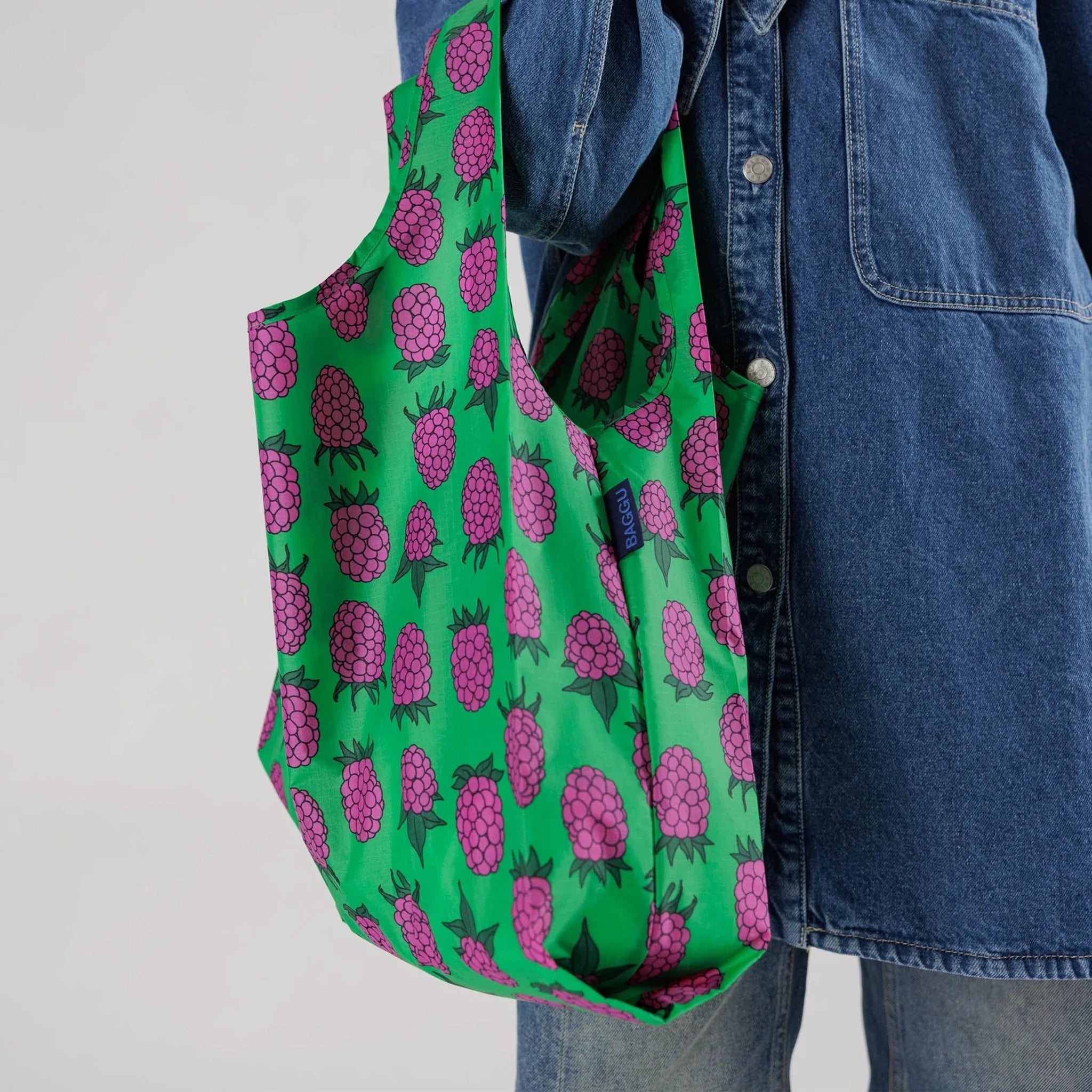 On a white background is a model holding a green reusable nylon tote bag with a purple ish pink raspberry pattern all over. 
