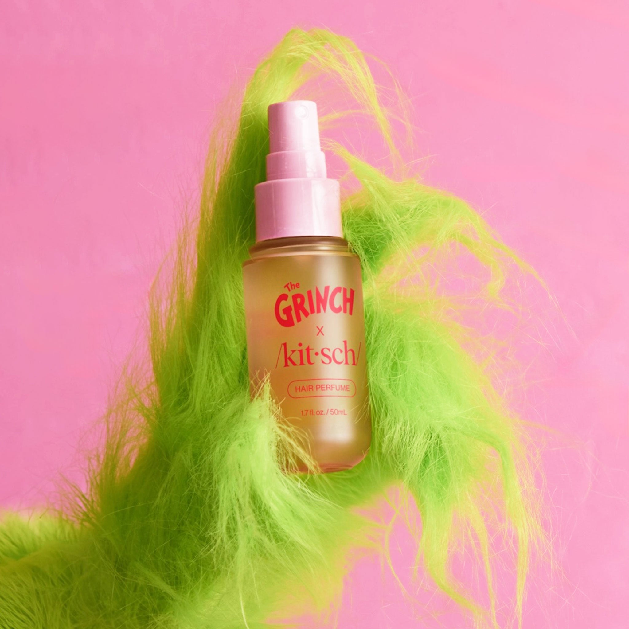 A frosted spray bottle labeled ‘The Grinch x Kitsch’ hair perfume is held upright in a shaggy bright-green hand against a pink background.