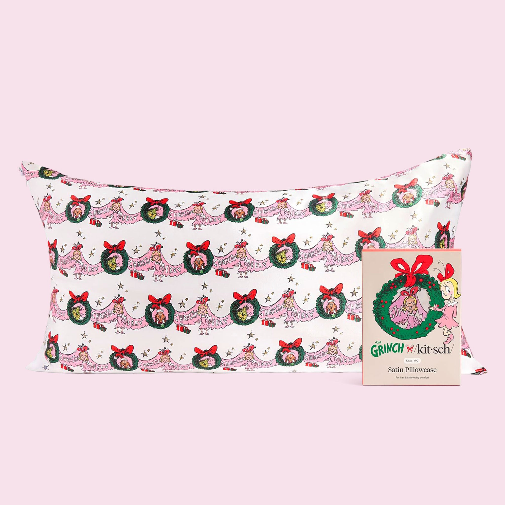 A white satin pillowcase printed with repeating Grinch holiday illustrations—wreaths with red bows and Cindy-Lou Who figures—shown on a pale pink background with its matching ‘The Grinch x Kitsch’ satin pillowcase box in front.