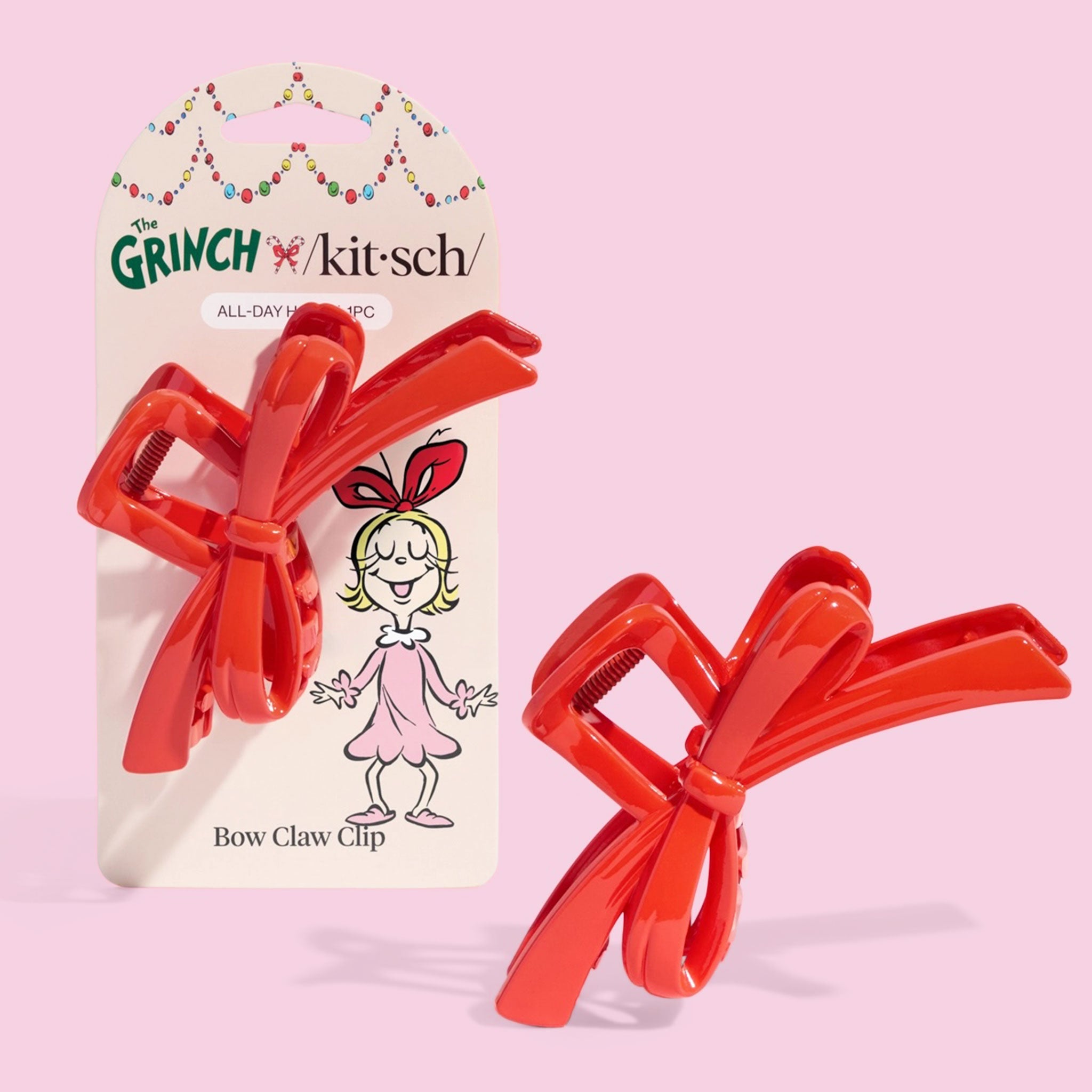 Two shiny red bow-shaped hair claw clips are shown in front of their ‘The Grinch x Kitsch’ packaging, which features a cartoon girl with a large bow, all on a light pink background.