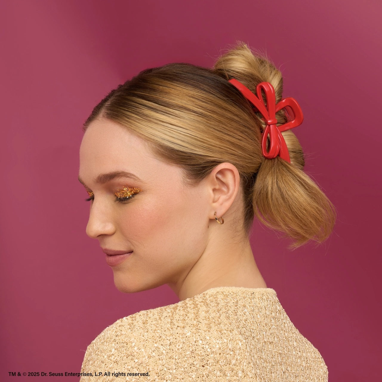 Side profile of a woman with blonde hair in a low bun, wearing a shiny red bow-shaped hair clip; she has gold glitter eye makeup and a sparkly beige top against a deep pink background
