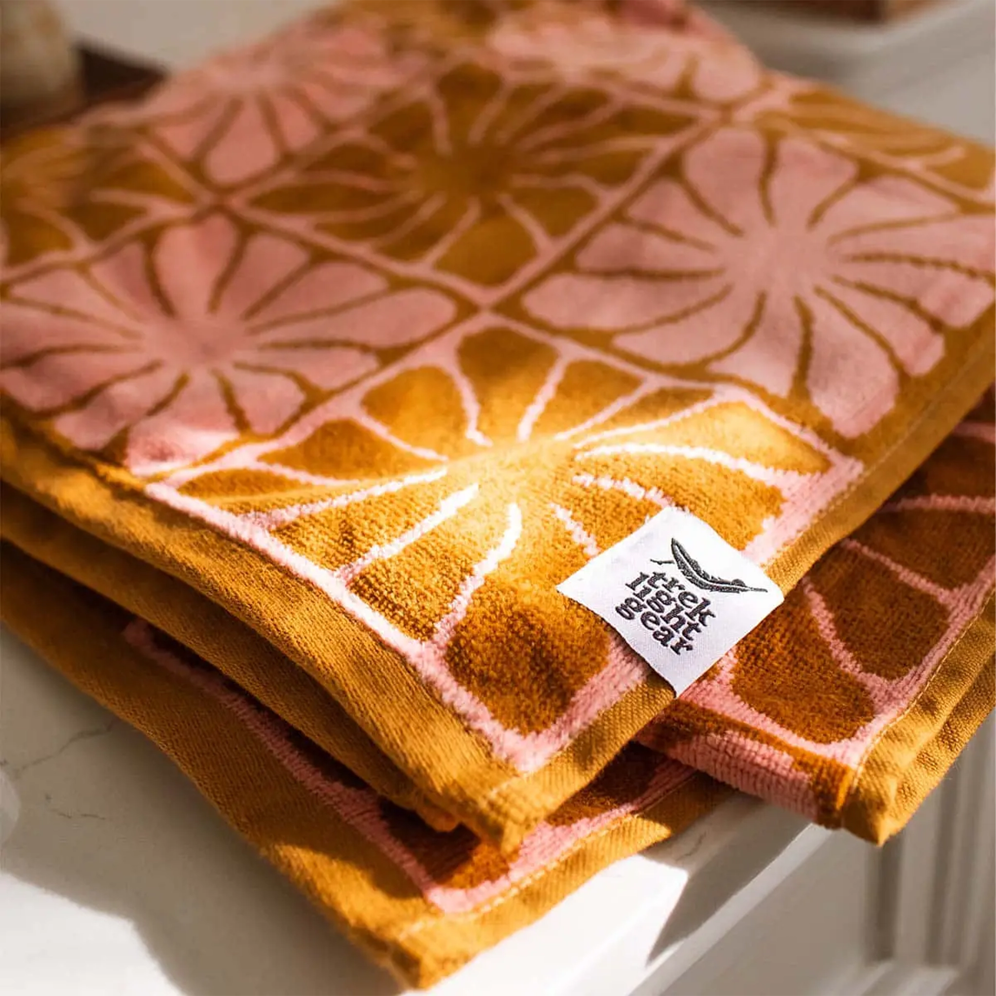 A pink and burnt orange bath/beach towel with a wavy floral checker design in each of the squares photographed here folded on a counter. 
