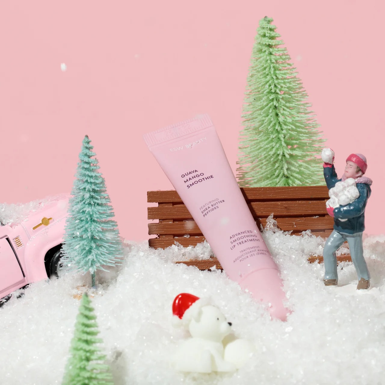 Guava Mango Smoothie lip treatment tube staged in a miniature winter scene with faux snow, small pine trees, a pink truck, and tiny figurines, against a pastel pink backdrop.