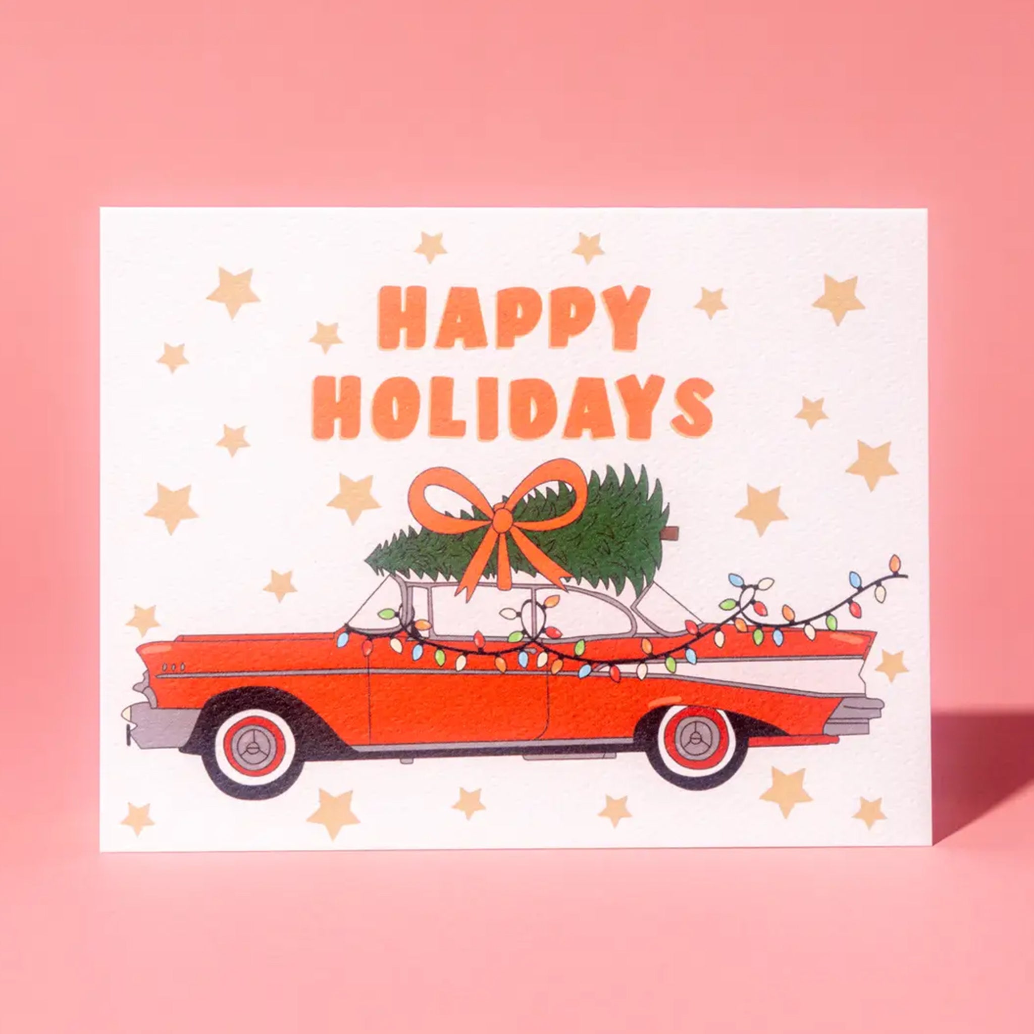 A white, red and green holiday card and envelope with a graphic of a vintage red car with a tree on top and text that reads, 'Happy Holidays'. 