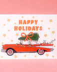 A white, red and green holiday card and envelope with a graphic of a vintage red car with a tree on top and text that reads, 'Happy Holidays'. 
