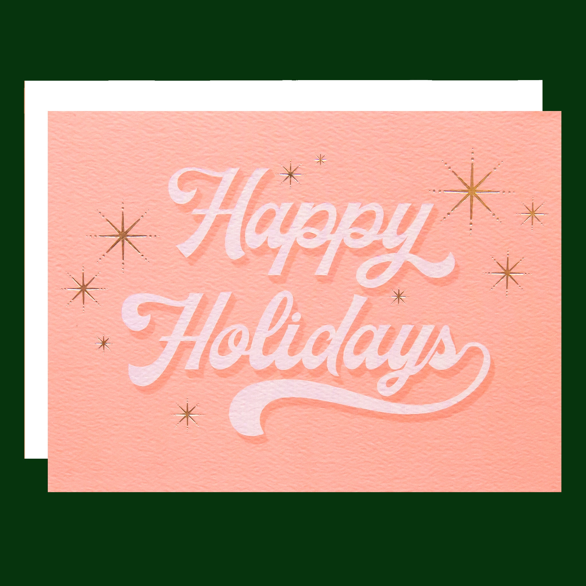 Pink greeting card with white cursive text that reads happy holidays surrounded by six gold foil starbursts. The card is accompanied by a solid white envelope.