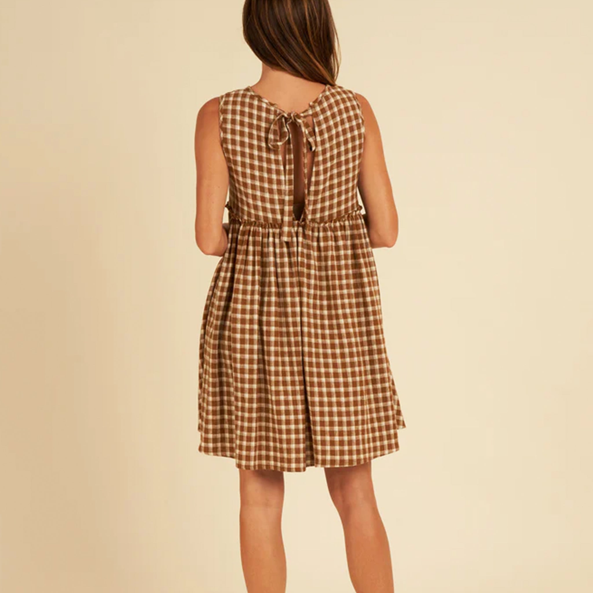 On a tan background is a model wearing a brown and tan plaid babydoll dress with a tie detail in the back. 