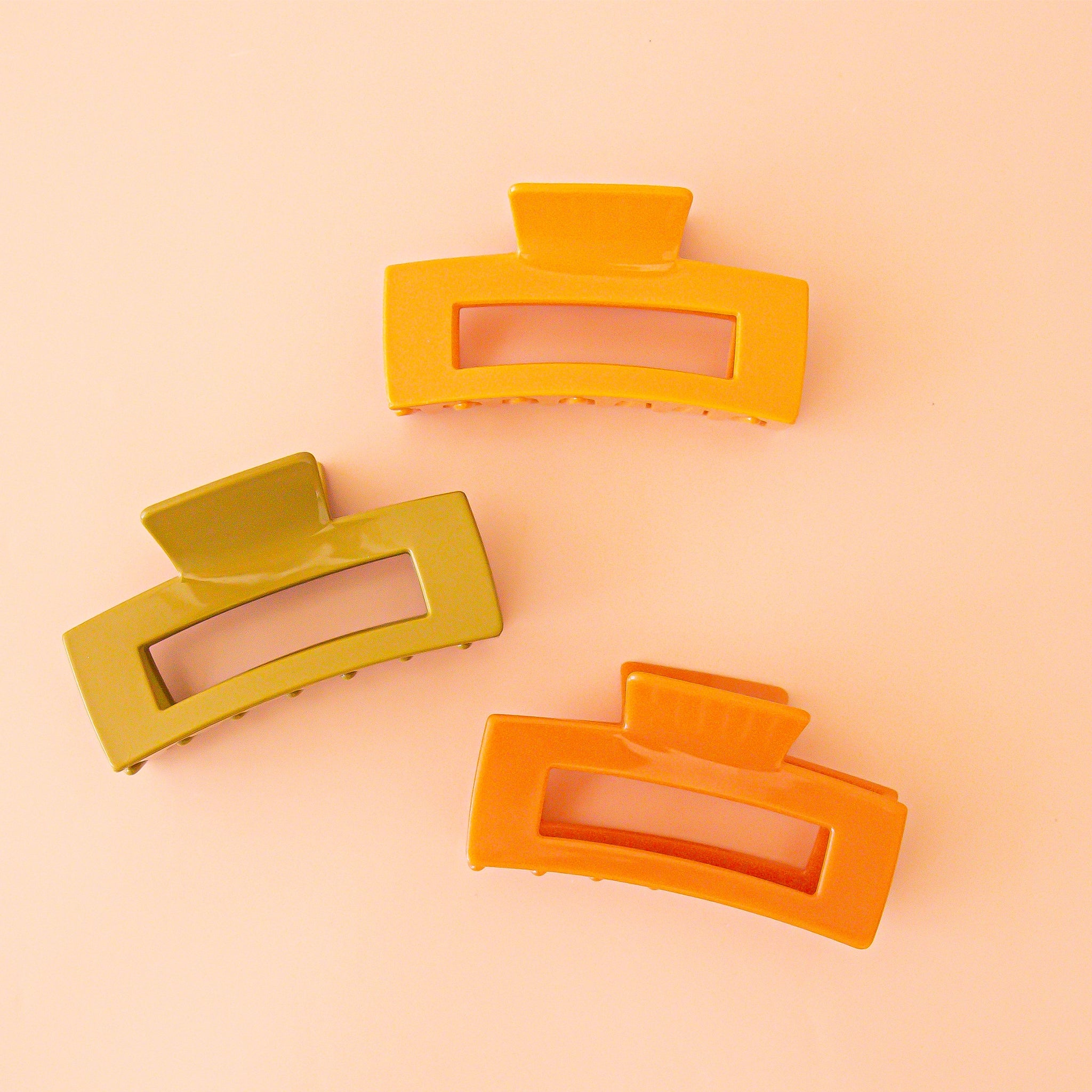 On a peachy background is three rectangle claw clips in different colors. One yellow, one olive, and one orange.