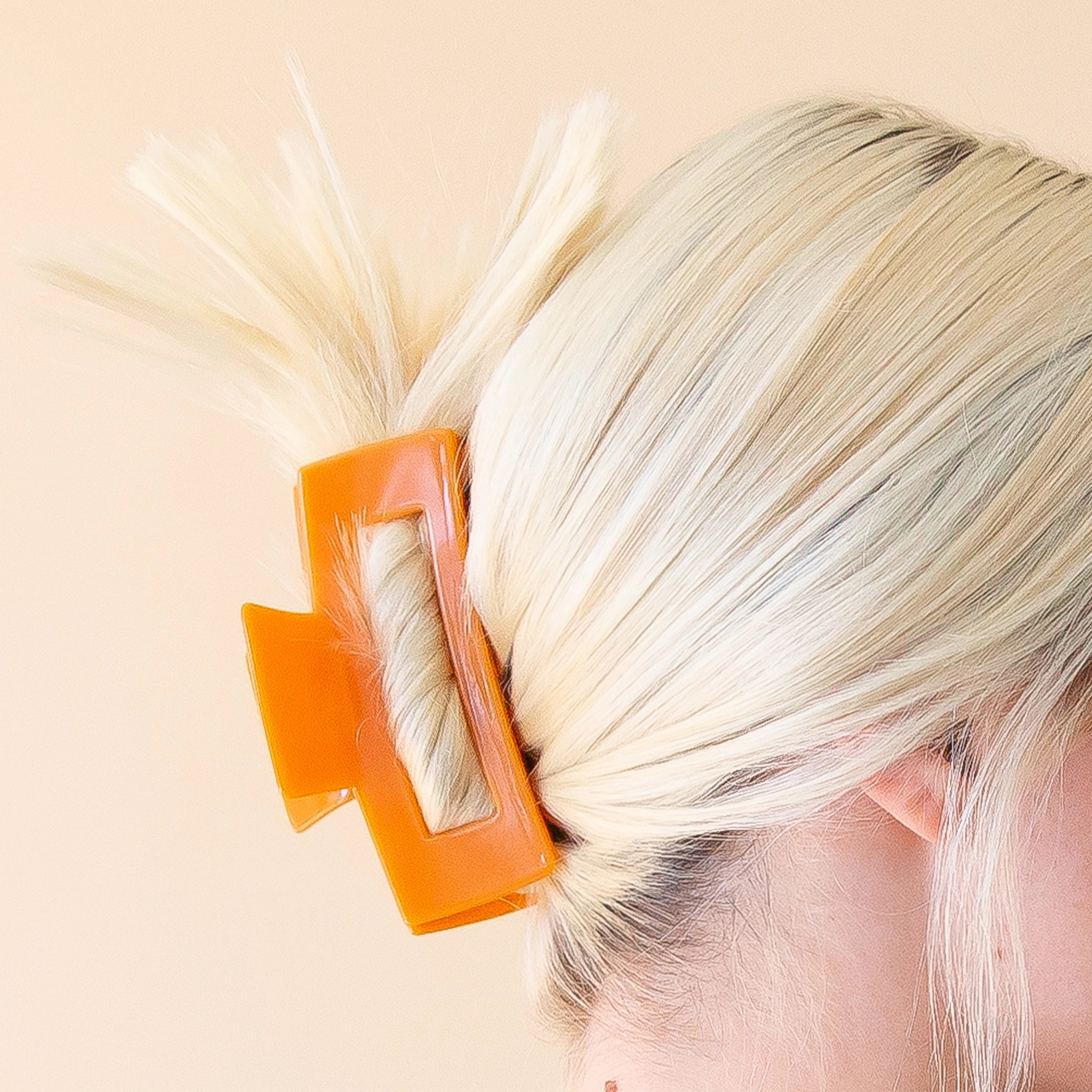 On a light peach background is a model wearing the Tangerine Havana Claw Clip that features a rectangular shape, a bright orange shade and a glossy finish. 