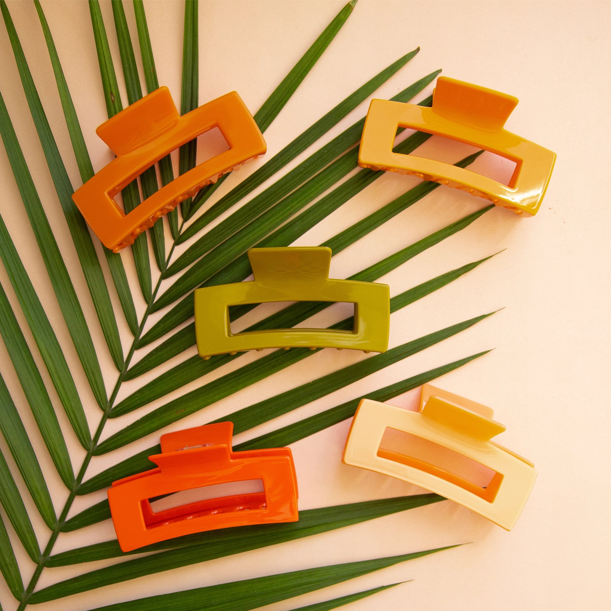 On a peachy background is a variety of rectangle claw clips in different colors on a green palm leaf.