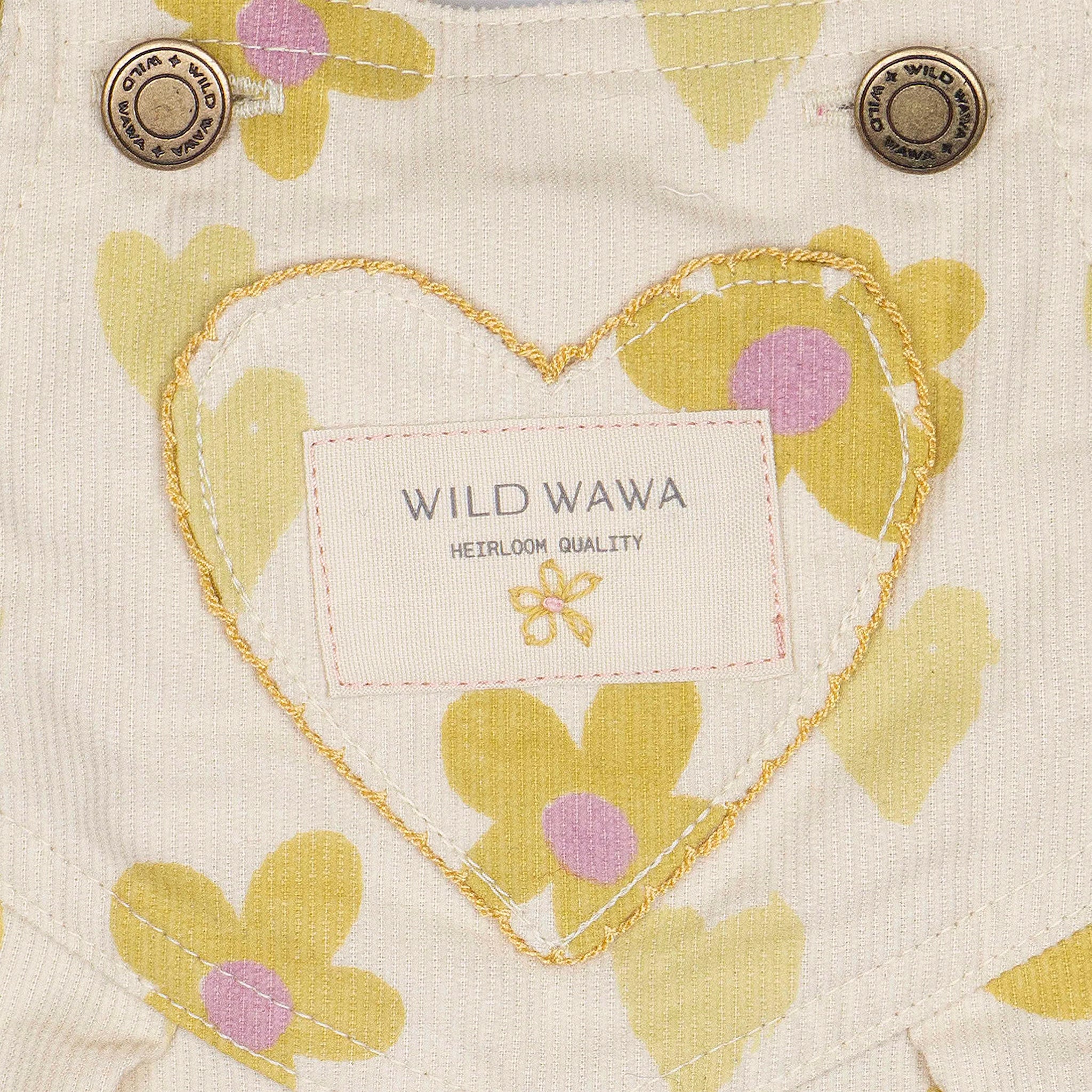 A cream colored pair of overalls with a yellow and pink floral and heart pattern. 
