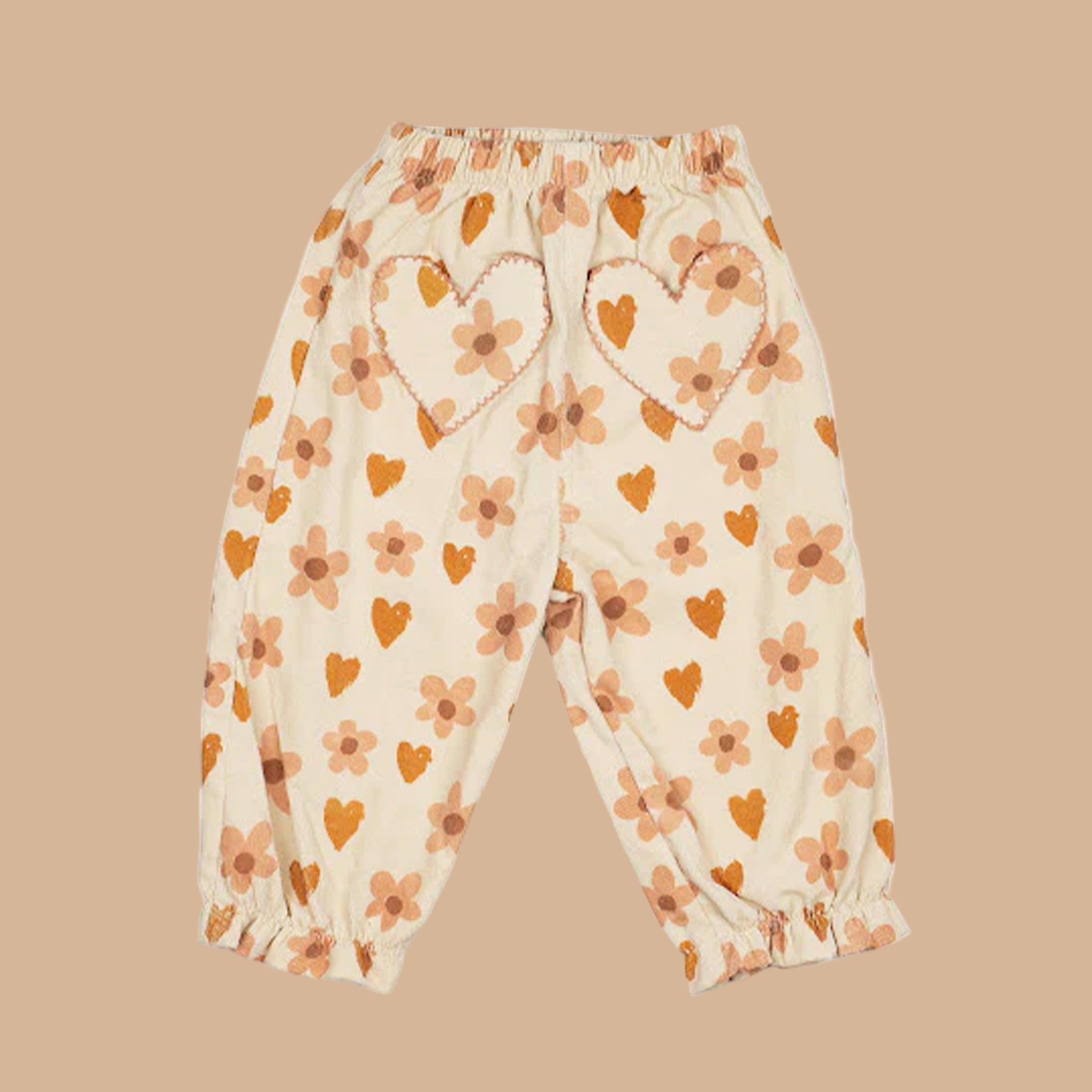A pair of neutral kids pants with an orange heart and floral print and heart shaped pockets. 